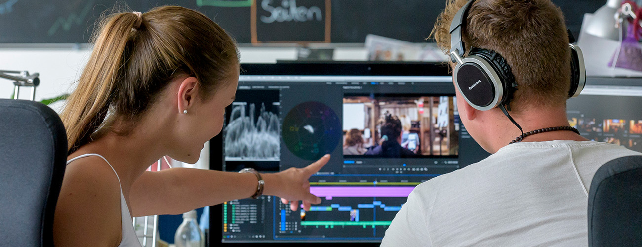 Ein Mann und eine Frau sitzen gemeinsam vor einem Monitor, auf dem ein Video geschnitten wird. Die Frau seigt mit ihrem Finger auf etwas in dem Video. Beide sind schräg von hinten fotografiert. - Quelle: WFB/Frank Pusch