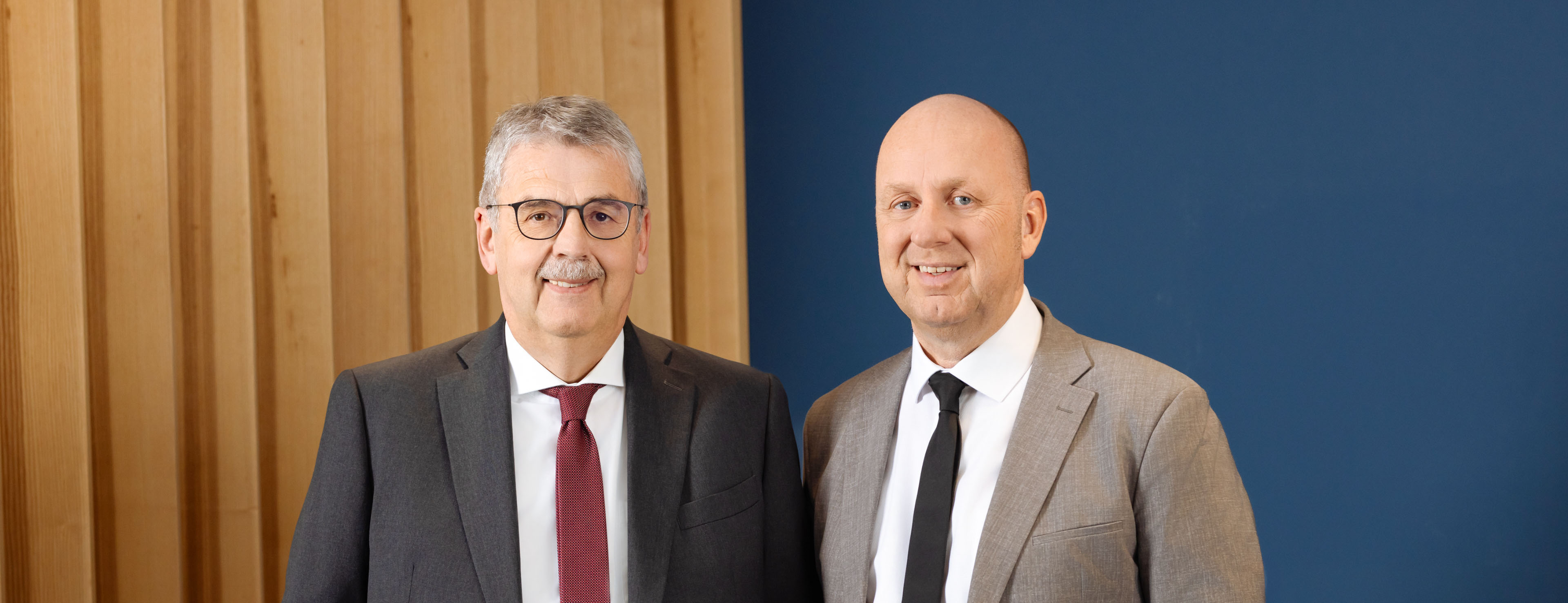 Zwei Männer in Anzügen stehen vor einem hellen Holzelement und einer dunkelblauen Wand. Sie lächeln. - Quelle: WFB / Jan Seebeck