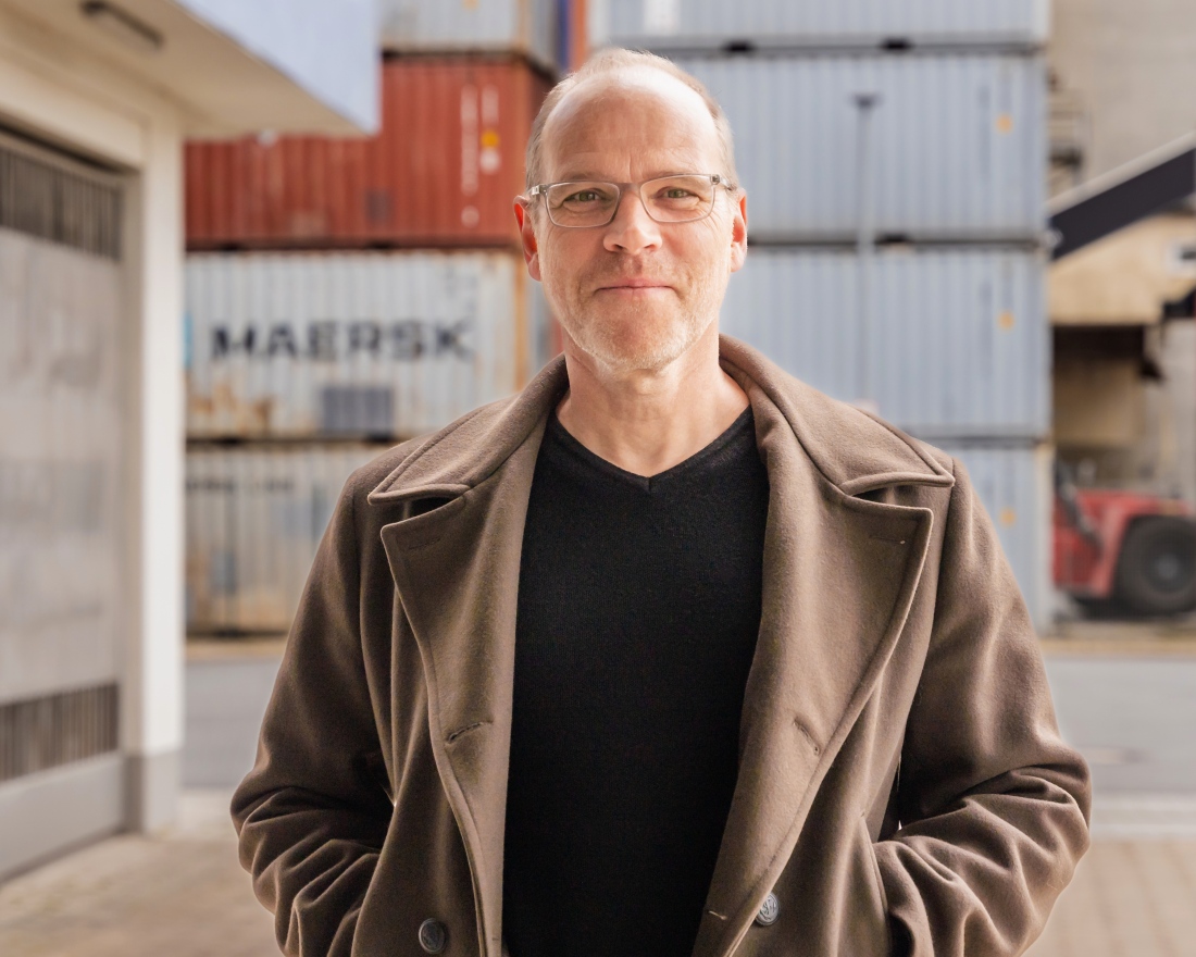 Ein Mann mit Brille blickt in die Kamera. Er steht in einem Containerhafen Ein Mann mit Brille blickt in die Kamera. Er steht in einem Containerhafen