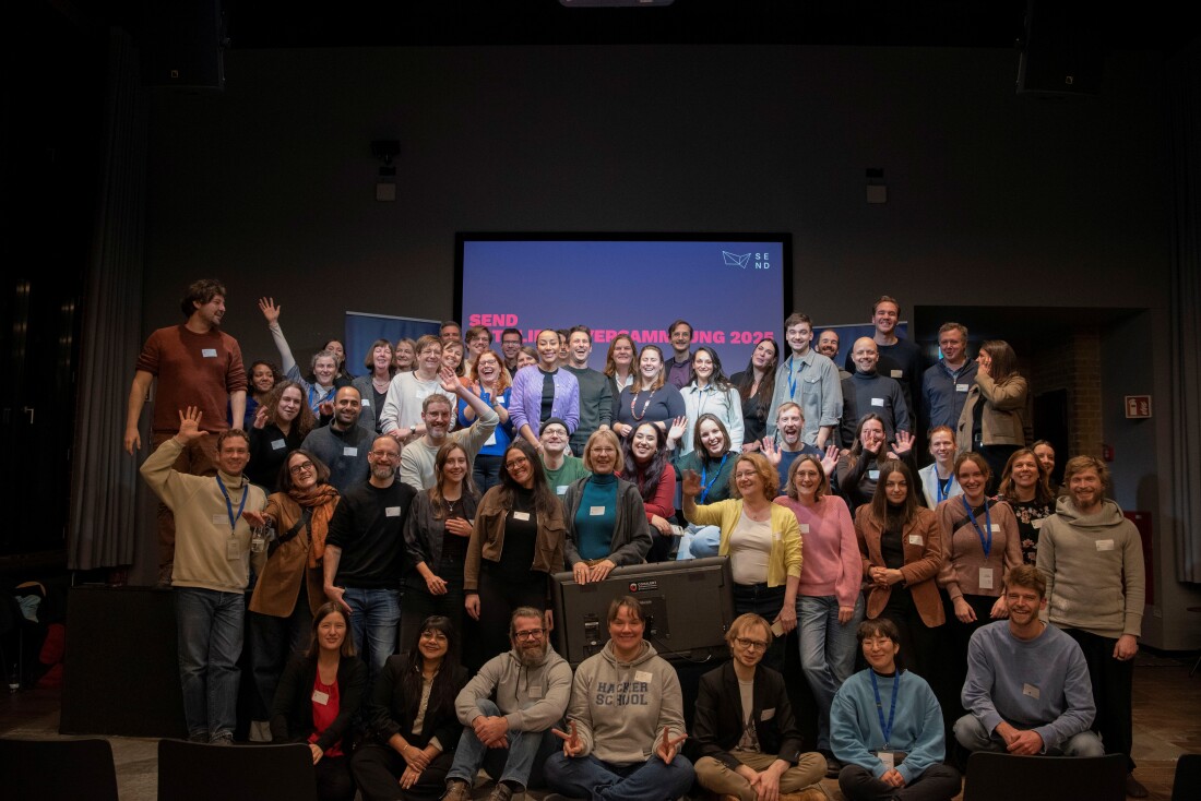 Eine Gruppe von 56 Personen sitzt oder steht auf dem Boden eines Raumes und schaut fröhlich in die Kamera. Im Hintergrund befindet sich ein Monitor mit der Aufschrift SEND.