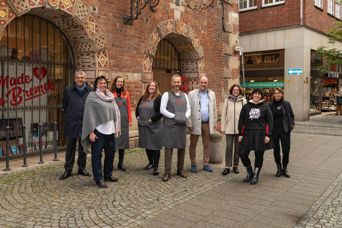 Fünf Gewinner:innen freuen sich über die Umsetzung der neuen Ideen in der Bremer Innenstadt.  Drei Männer und sechs Frauen stehen lächelnd vor einem sehr alten Gebäude, auf einem der Schaufenster steht "Made in Bremen", im Hintergrund ist eine Buchhandlung zu sehen.