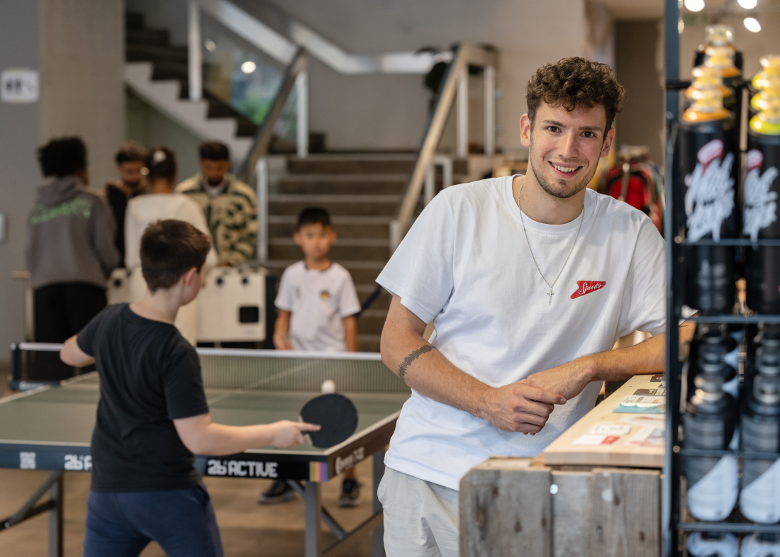 Ein junger Mann lehnt lächelnd an einem Holztresen, im Hintergrund spielen zwei Kinder Tischtennis.