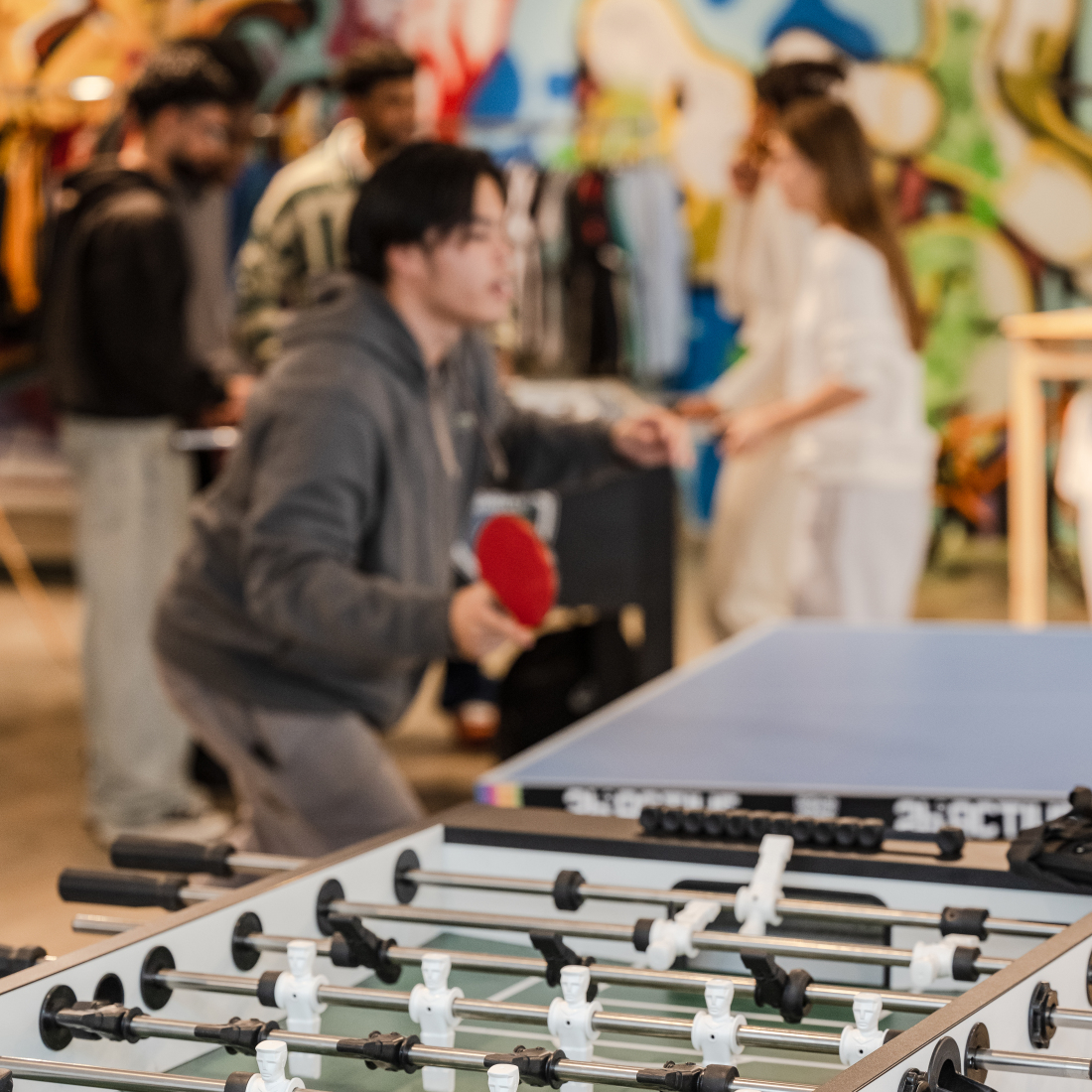 Ein junger Mensch spielt an einem Ende einer Tischtennisplatte. Im Hintergrund stehen vier Personen an einem Tischkicker.