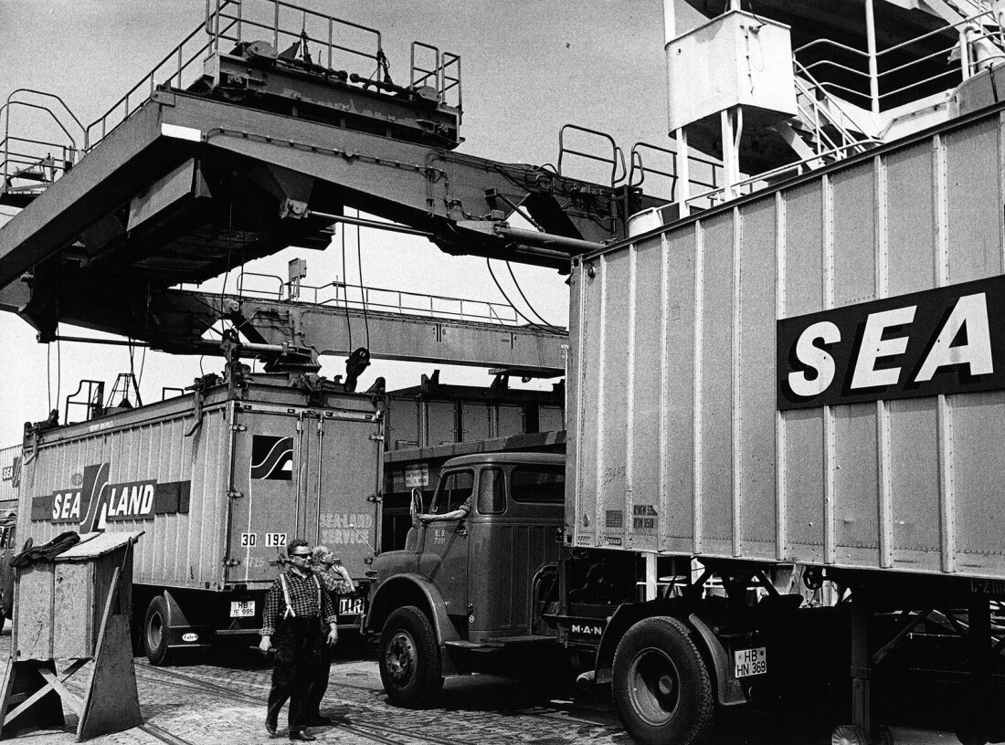 Schwarz-Weiß-Aufnahem aus den 60-er Jahren: Zwei Männer vor Lastwagen, die unter Kränen stehen, die Container verladen.