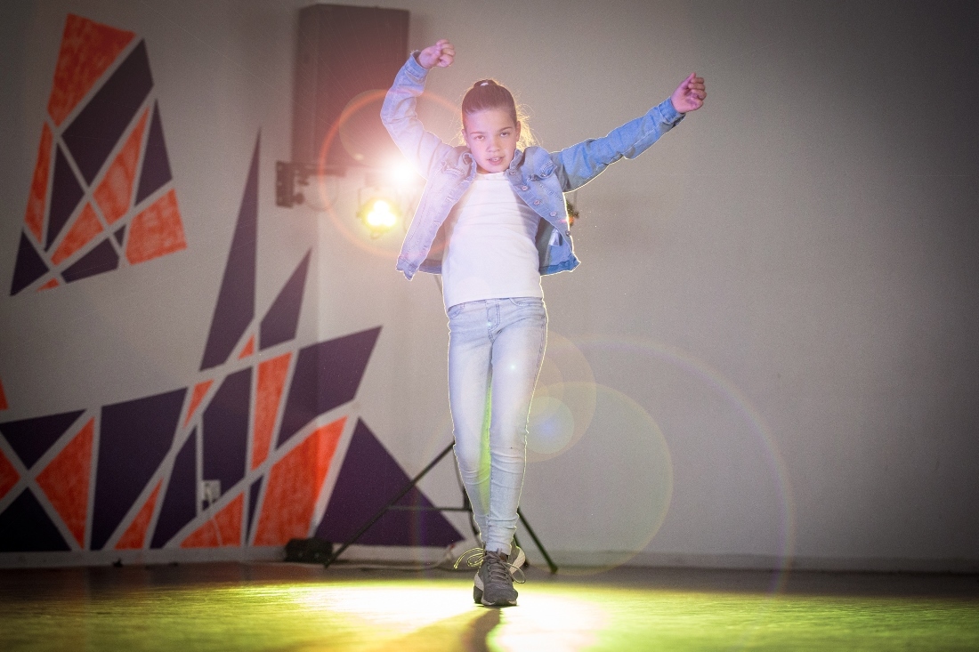 Ein zehnjähriges Mädchen in Turnschuhen, Jeans, weißem Shirt und Jeansjacke tanzt vor einem Scheinwerfer. Sie springt hoch und wirft ihre Arme in die Luft.