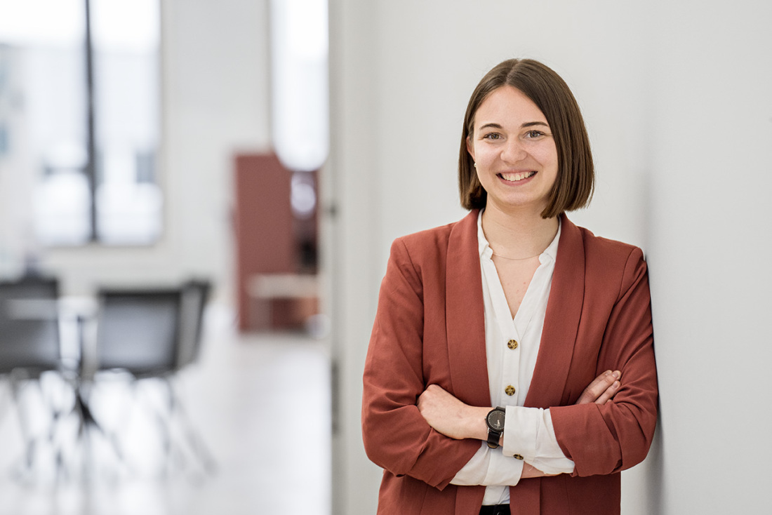 Eine Frau mit braunen Haaren und einem braunen Blaser lehnt sich an eine Wand