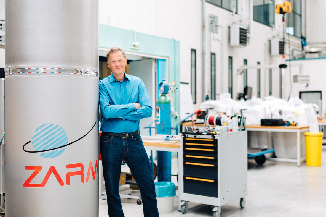 Ein Mann in blauem Hemd und Jeans steht lächelnd mit verschränkten Armen an eine Säule mit dem Schriftzug "ZARM" gelehnt. Im Hintergrund eine große Halle mit technischer Ausrüstung auf Tischen und Rollwägen..