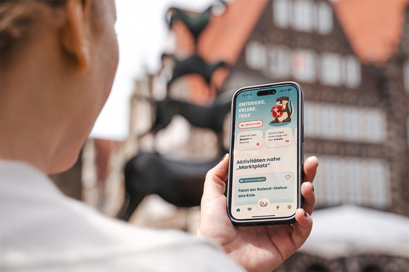Eine Hand hält ein Smartphone. Auf dem Display ist eine App geöffnet, mit der man Erlebnisse in Bremen finden kann.