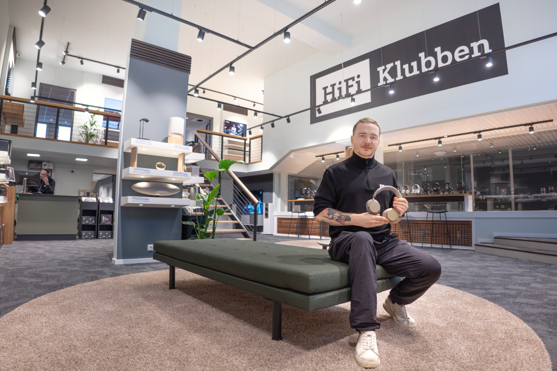 Ein Mann sitzt in einem Laden auf einem grünen Hocker und hält Kopfhörer in der Hand. Im Hintergrund das Logo von HiFi Klubben