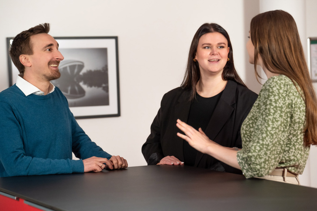 Zwei Frauen und ein Mann stehen um einen Tisch herum und unterhalten sich