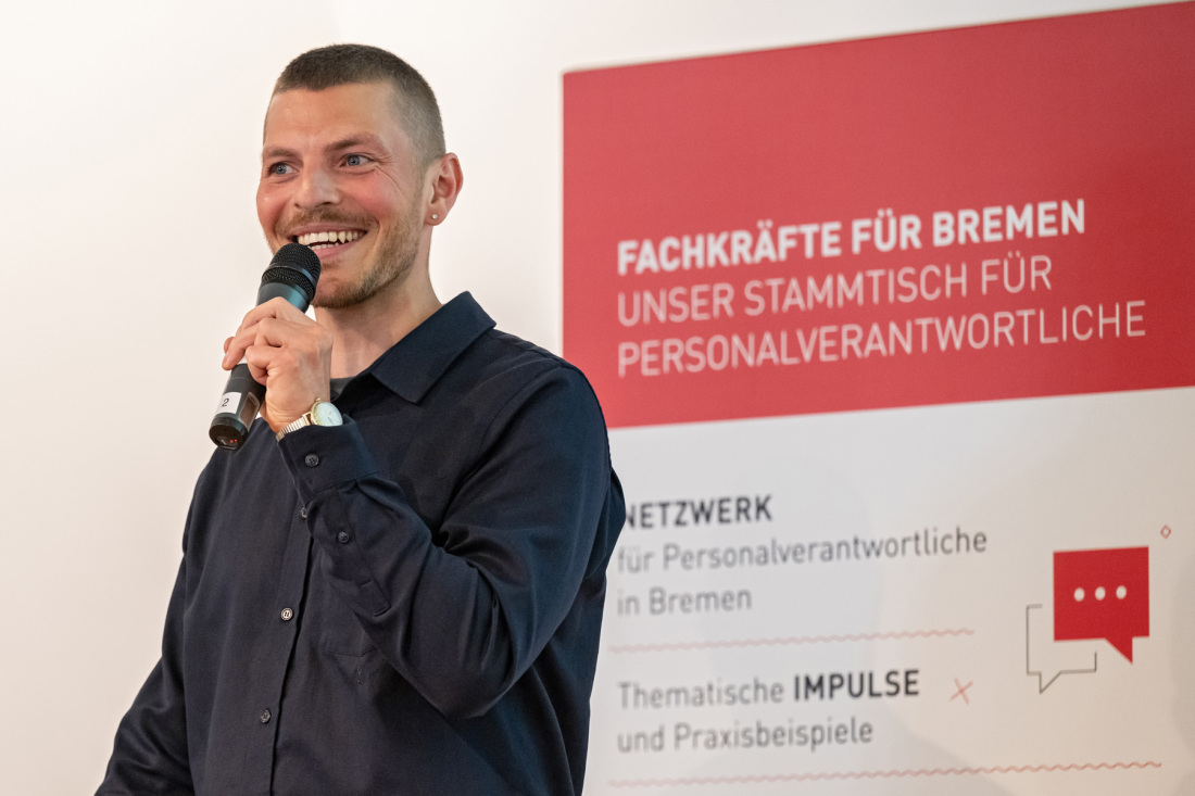 Ein Mann spricht in ein Mikro. Hinter ihm ein Roll-Up mit der Aufschrift Stammtisch für Personalverantwortliche Ein Mann spricht in ein Mikro. Hinter ihm ein Roll-Up mit der Aufschrift Stammtisch für Personalverantwortliche