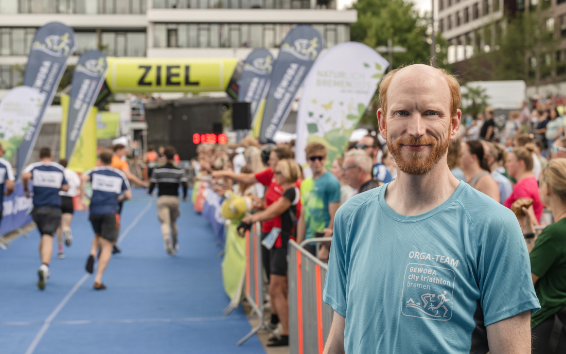 Ein Mann, Jan Brüning, steht im Vordergrund, wärhend im Hintergrund Menschen in ein Ziel einlaufen Ein Mann, Jan Brüning, steht im Vordergrund, wärhend im Hintergrund Menschen in ein Ziel einlaufen