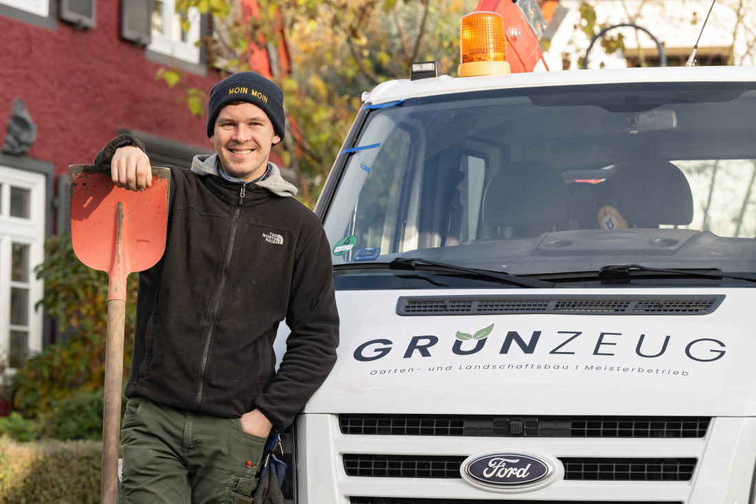 Ein junger Mann steht vor einem weißen Fahrzeug. Auf dem Fahrzeug steht Grünzeug