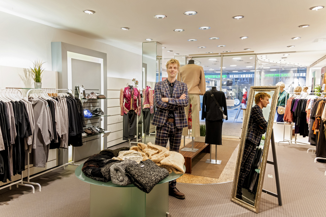 Ein blonder Mann steht lächelnd in einer Lädenfläche, um ihn herum Kleiderständer und Tische mit Mode.