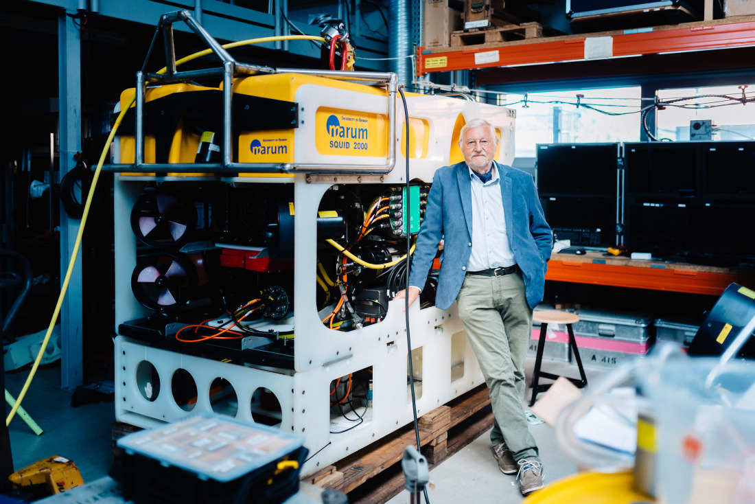 Gerold Wefer, ein Mann mit weißen Haaren, Brille und kurzem Bart, lehnt sich an einen mannshohen eckigen Forschungsapparat, auf dem das Logo des MARUM – Zentrum für Marine Umweltwissenschaften zu sehen ist.