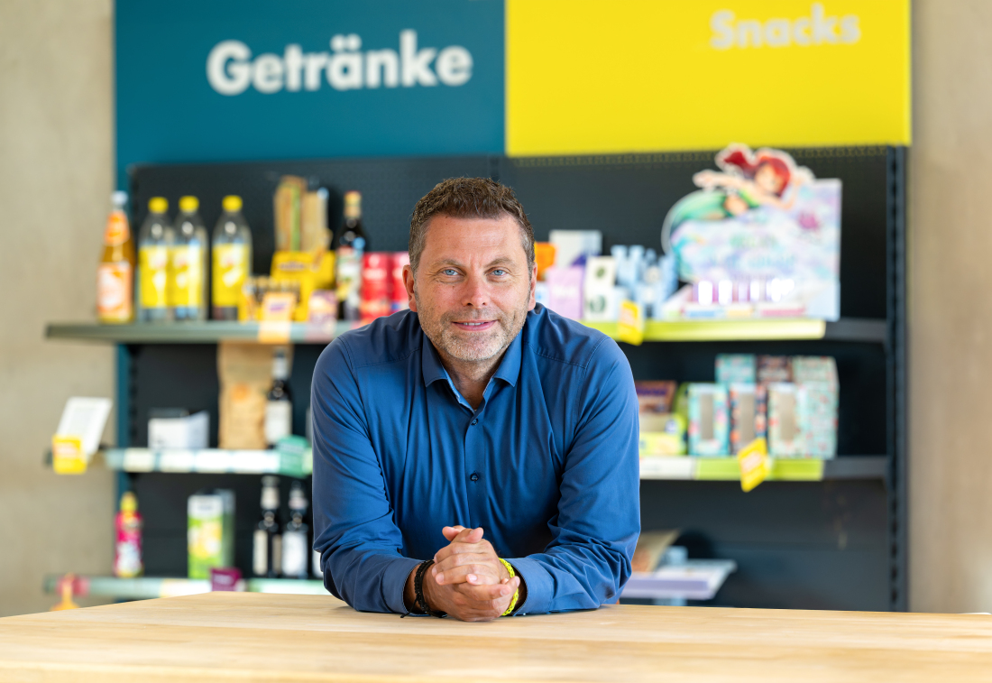 „Ein Mann in blauem Hemd lehnt an einem Holztisch und blickt in die Kamera. Hinter ihm stehen Regale mit Getränken, Snacks und weiteren Produkten. „Ein Mann in blauem Hemd lehnt an einem Holztisch und blickt in die Kamera. Hinter ihm stehen Regale mit Getränken, Snacks und weiteren Produkten.