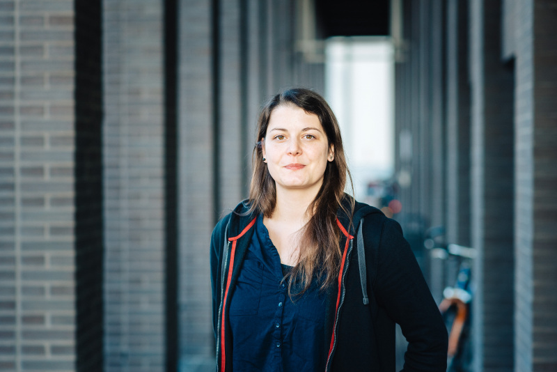 Eine junge Wissenschaftlerin mit langem Haar steht in einem Gebäudeflur vor Backsteinsäulen
