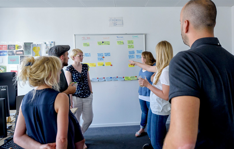 Eine Gruppe Menschen steht vor einem Whiteboard. Sie schauen auf viele bunte Klebezettel und diskutieren darüber.