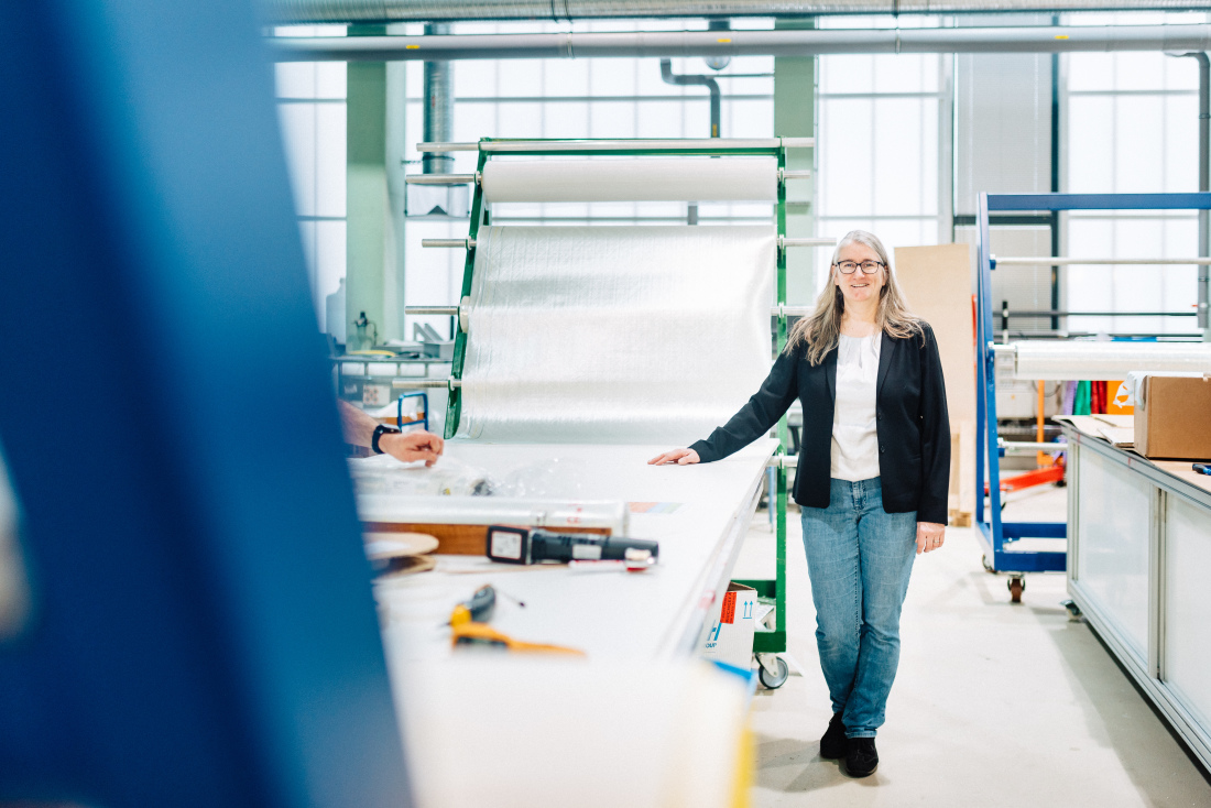 Eine Frau mit langen Haaren und schwarzer Brille steht in einer Halle, die rechte Hand liegt auf einem Werktisch, auf dem ein dünnes Material ausgebreitet ist.