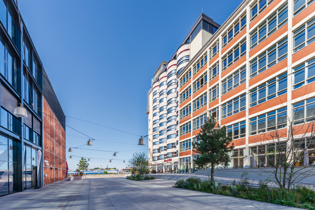 Ein weitläufiger Plattenweg zwischen den modernen Gebäuden John & Will Hotel auf der rechten Seite und der Brüning Group auf der linken Seite. An das Hotel schließt sich eine Gastronomie an.