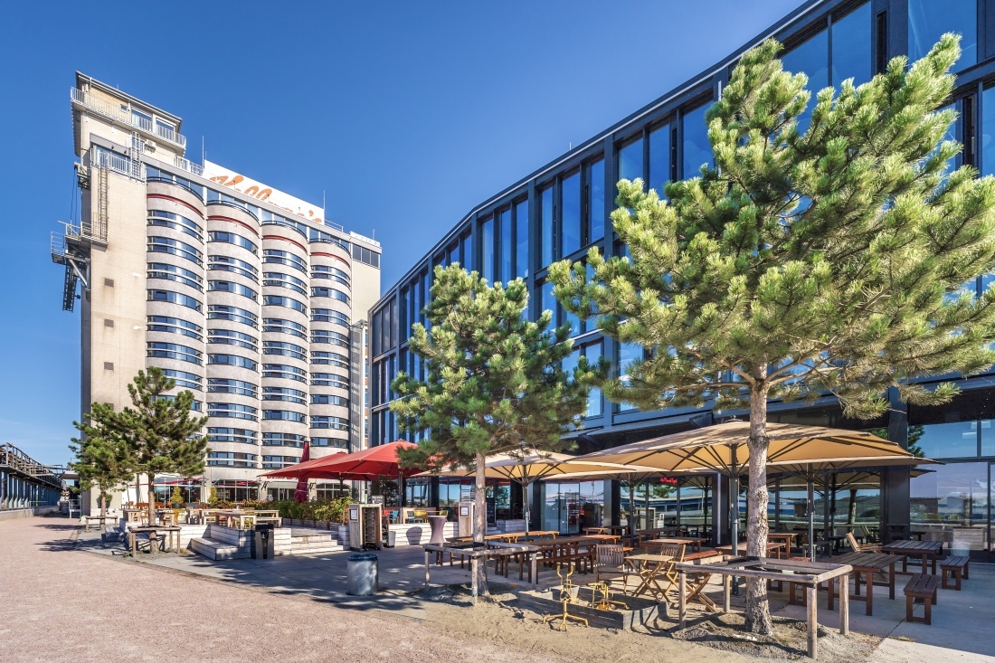 Im Vordergrund eine Gastronomie mit einer Terasse, Sonnenschirmen und Bäumen. Im Hintergrund ragt das John & Will Silo-Hotel in den blauen Himmel Im Vordergrund eine Gastronomie mit einer Terasse, Sonnenschirmen und Bäumen. Im Hintergrund ragt das John & Will Silo-Hotel in den blauen Himmel