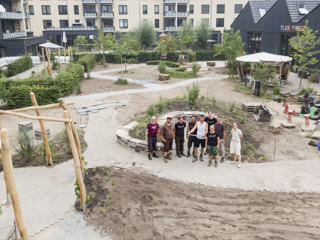 Eine Gruppe Menschen steht in einem neu angelegten Garten in einem Wohngebiet. Sie sind umgeben von Wohnhäusern.