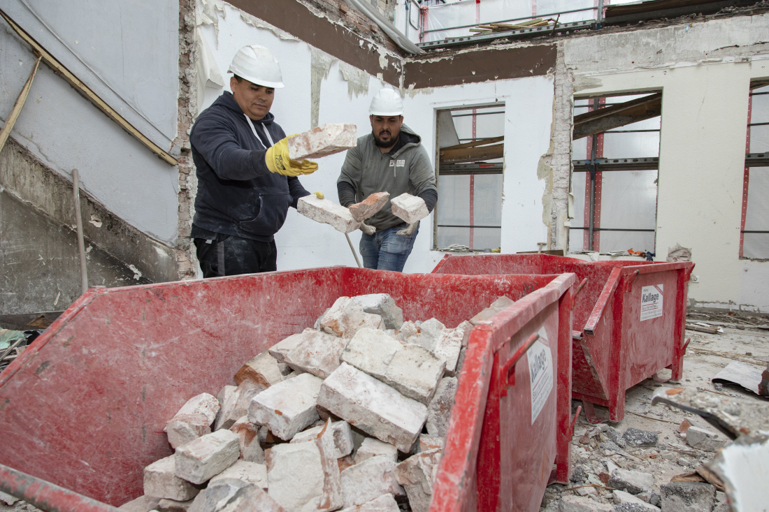 Zwei Männer werfen Ziegelsteinen in einen roten Container auf einer Baustelle