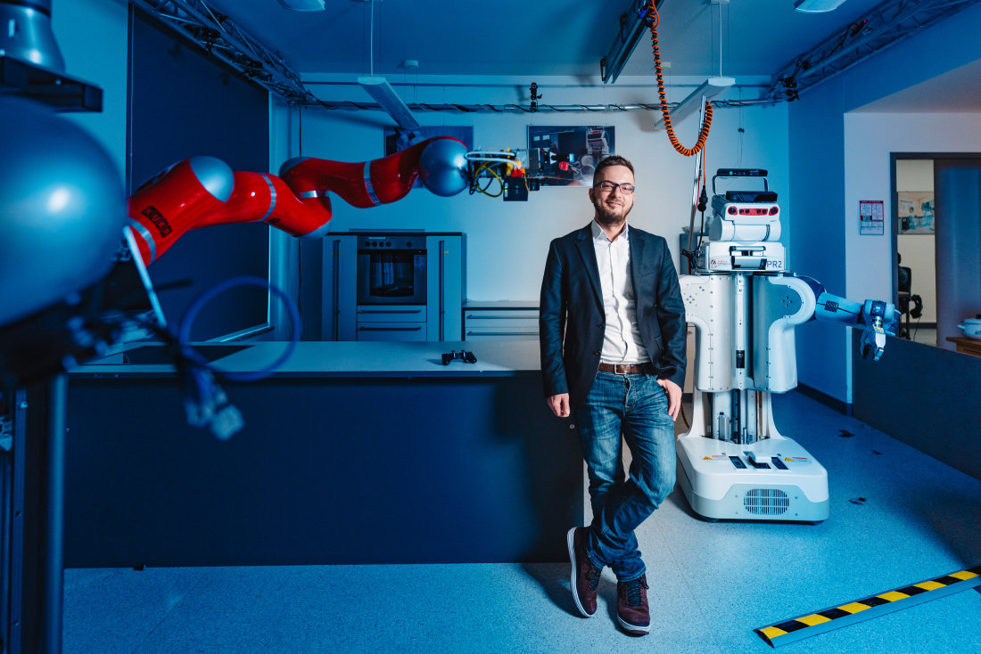 Ein Mann mit Brille in Jeans und Jacket steht in einem bläulich beleuchteten Raum und lächelt. Links von ihm ist ein langer roter Roboterarm zu sehen, rechts von ihm steht ein mannsgroßer weißer Roboter im Hintergrund.