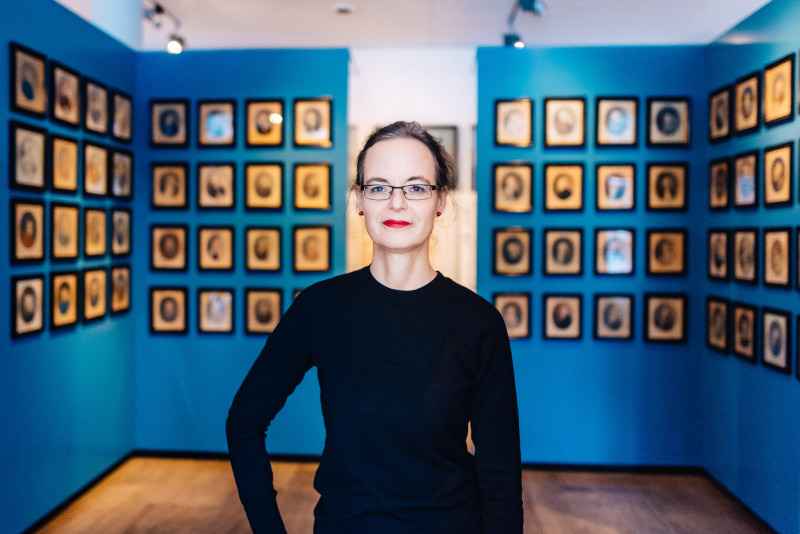 Eine Frau mit Brille steht lächelnd vor einer blauen Wand mit vielen Bilderrahmen.