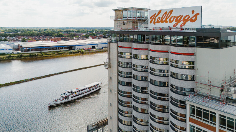 Das Bild zeigt ein hohes Gebäude mit dem Kellogg’s-Schriftzug am Wasser. Ein Ausflugsschiff fährt auf dem Fluss daneben vorbei.