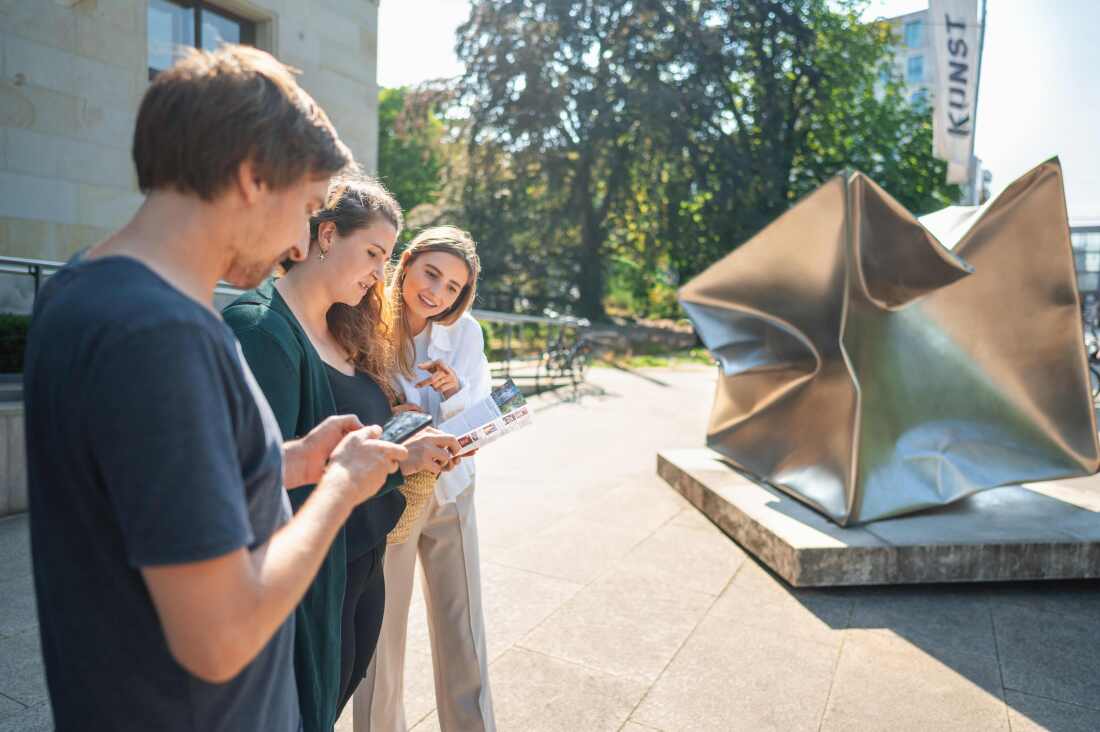 Drei junge Menschen stehen neben einem Kunstwerk und blicken auf Infomaterial Drei junge Menschen stehen neben einem Kunstwerk und blicken auf Infomaterial
