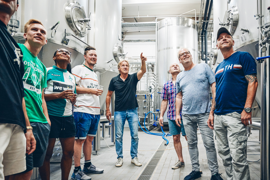 Eine Gruppe von acht Personen steht in einer Brauerei zwischen großen Edelstahltanks. Ein Mann in der Mitte erklärt und zeigt nach oben, während die anderen interessiert zuhören und teils Getränke in der Hand halten. Eine Gruppe von acht Personen steht in einer Brauerei zwischen großen Edelstahltanks. Ein Mann in der Mitte erklärt und zeigt nach oben, während die anderen interessiert zuhören und teils Getränke in der Hand halten.