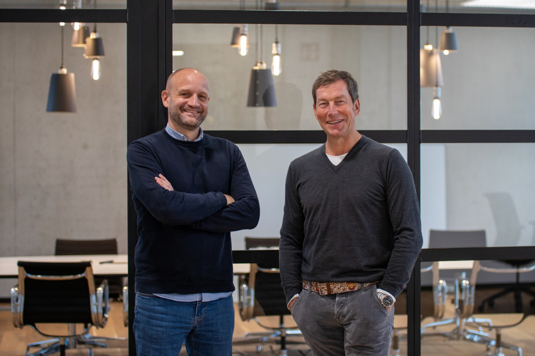 Two men stand in front of a conference room and smile into the camera