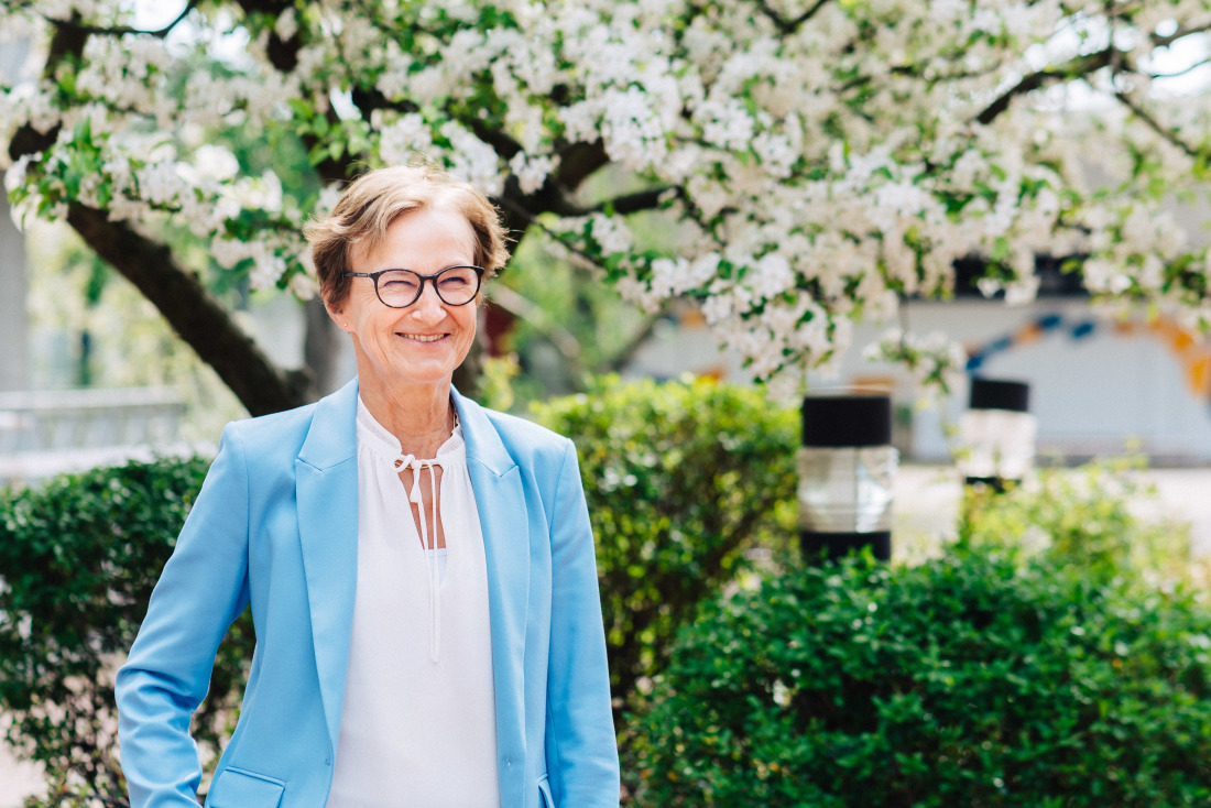 Eine Frau mit kurzen Haaren und schwarzer Brille steht lächelnd vor einem einem weiß blühenden Baum. Sie trägt einen hellblauen Blazer und eine weiße Bluse.