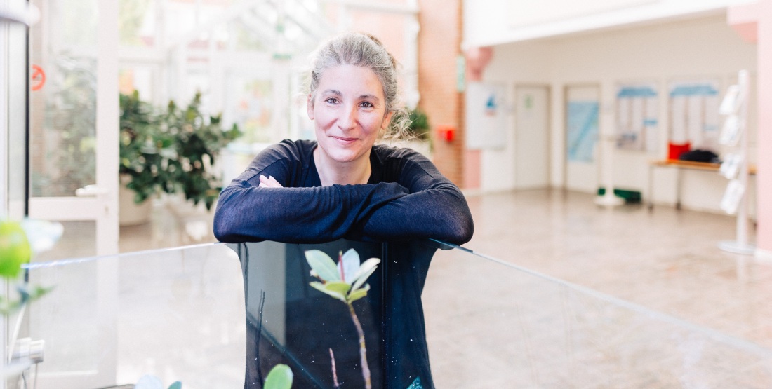 Eine Frau mit zurückgesteckten grauen Haaren lächelt in die Kamera. Sie steht in einem großen, hellen Raum und stützt sich auf einen brusthohen Glaskasten, in dem sich Pflanzen befinden.