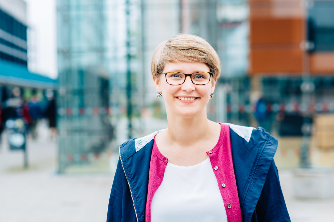 Eine junge Frau mit Brille steht lächelnd vor einem Gebäude.